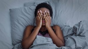 A woman covers her face while laying in bed. This could represent the anxious insomnia that online anxiety therapy in Chicago, IL can help with! Search "anxiety therapist near me" to learn of our services in Texas, Florida, and more! 60604